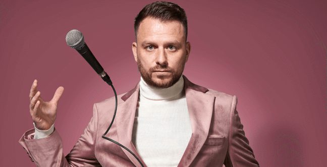 A man in a blush pink suit and white turtleneck throws his microphone in the air