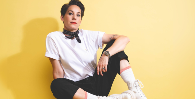 Suzi Ruffel sits with one leg up and her arm on top of her leg. She is wearing a white t-shirt with black trousers and a neckerchief round her neck