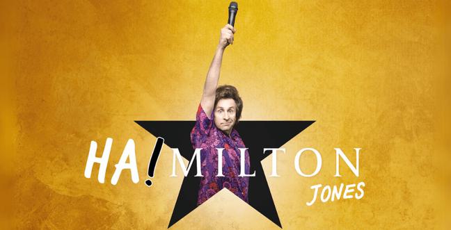 Milton Jones stands in a black star holding a microphone in the air with a gold backdrop