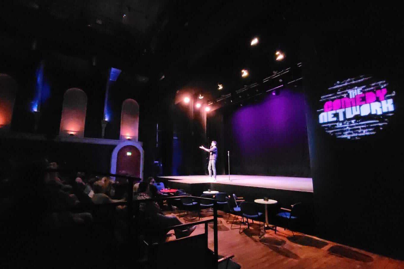 A comedian on stage with 'The Comedy Network' logo to the right of the stage, projected on to the wall