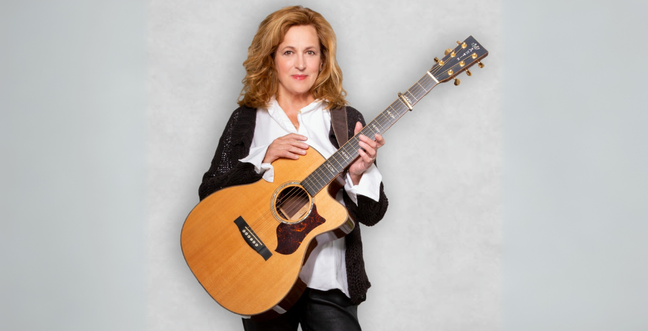 A woman stood on a white background, she is holding an acoustic guitar