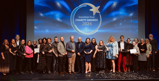 A selection of people dressed smartly stand on the stage smiling with Greenham Trust Charity Awards 2024 projected behind them