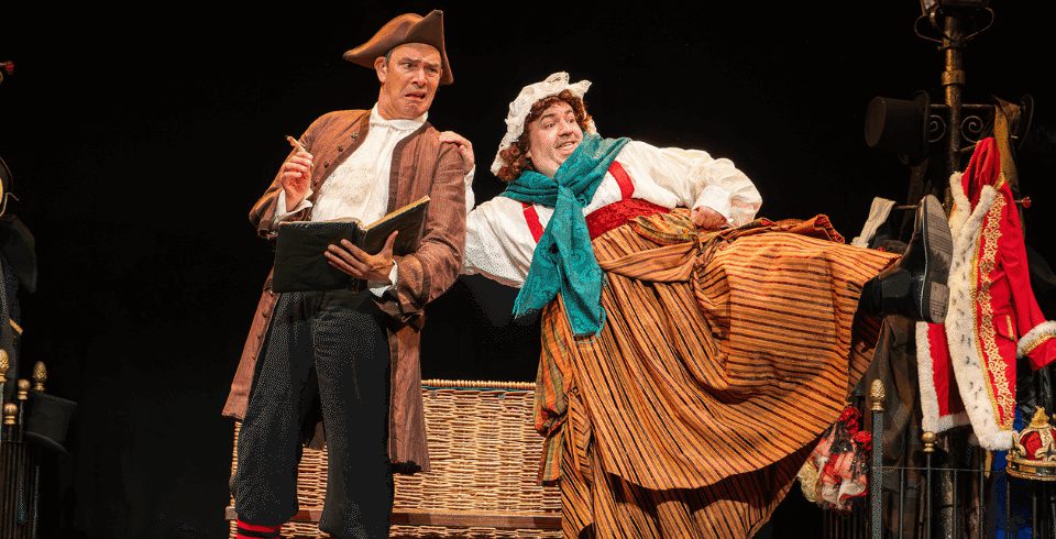 Two people on stage, one with a pirate hat holding a book and one with a person in a large skirt, green scarf and a bonnet