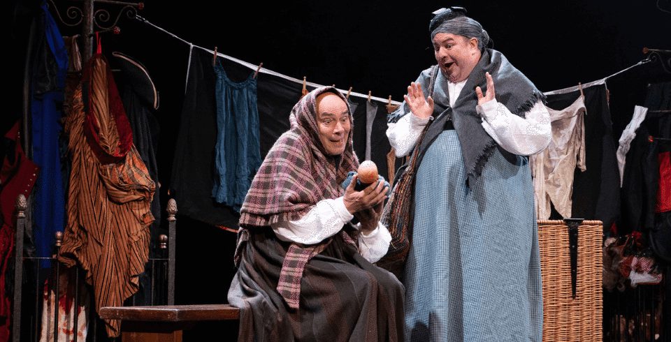 Two people on stage look at an apple. One person wears a shawl while holding an apple, while the other looks surprised wearing a grey shawl