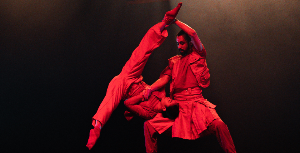 A man holds a woman against his thigh as she pushes her shoulder into his leg, allowing her to do the splits upside down