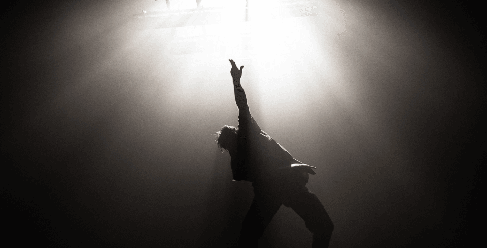 A dancer moves around a dark stage lit by one bright white light
