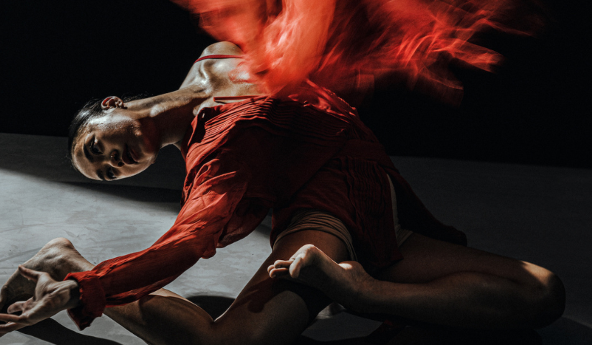 A woman in a red flowing outfits sits on the floor, leaning back. A red blur, almost like fire sits above her chair.