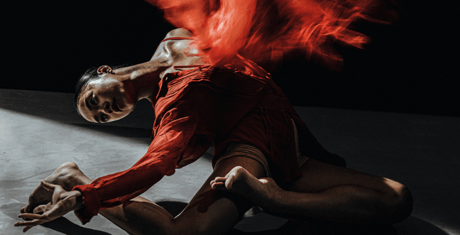 A woman in a red flowing outfits sits on the floor, leaning back. A red blur, almost like fire sits above her chair.