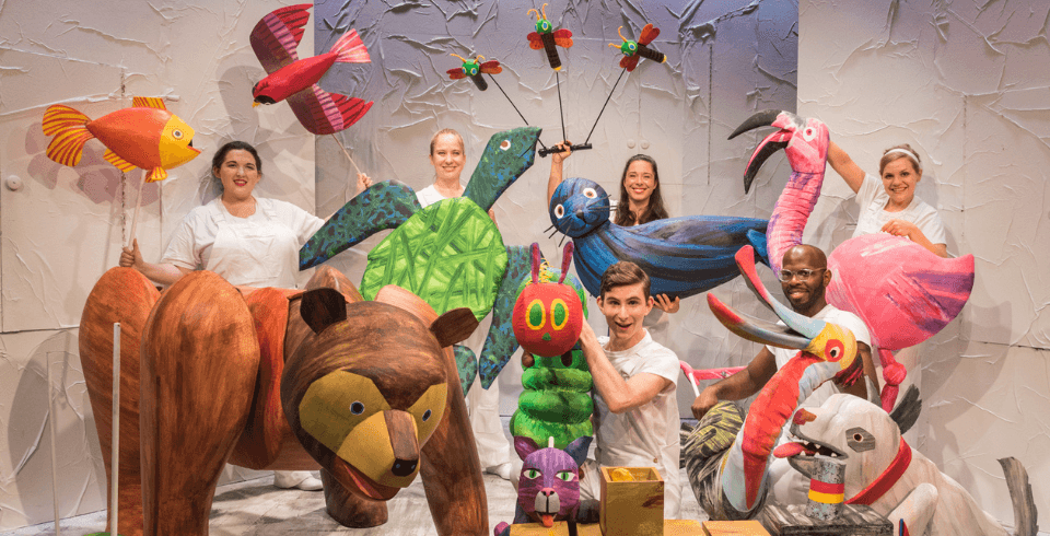 An array of actors dressed in white hold colourful puppets of various animals and insects