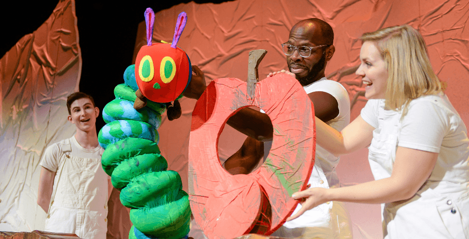 A man and a woman dressed all in white hold a giant green and red caterpillar puppet and a large red apple with a hole in the middle