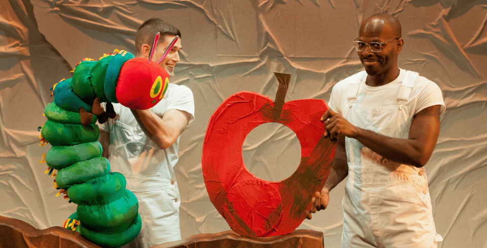 Two men dressed all in white hold a giant green and red caterpillar puppet and a large red apple with a hole in the middle