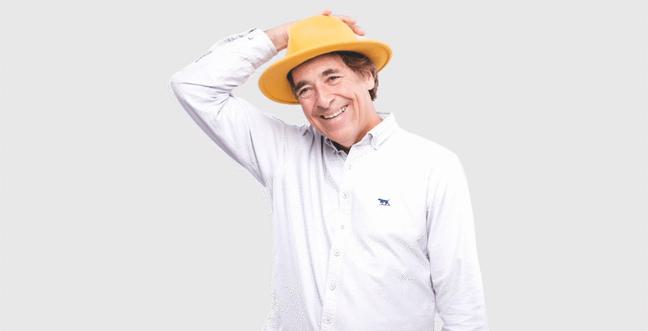 A man in a yellow hat wears a white shirt and a beige background. He is standing with one hand on his hat and smiling.