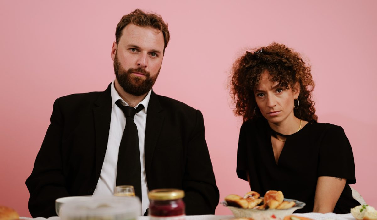 A man in a suit and woman in a black dress sit against a pink backdrop