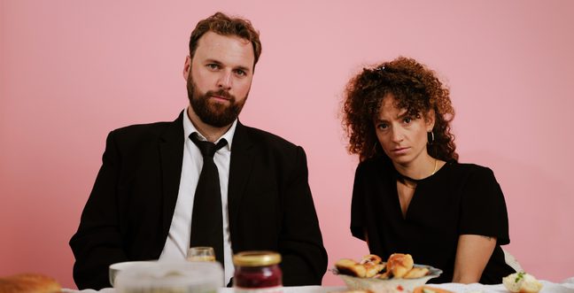 A man in a suit and woman in a black dress sit against a pink backdrop