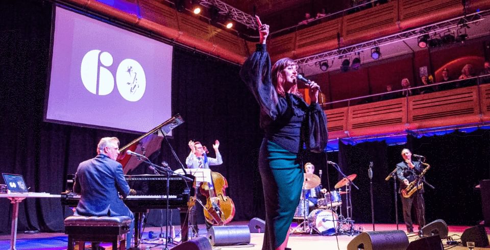 A jazz band on stage in the spotlight with a screen projected behind them with the number 60 on it