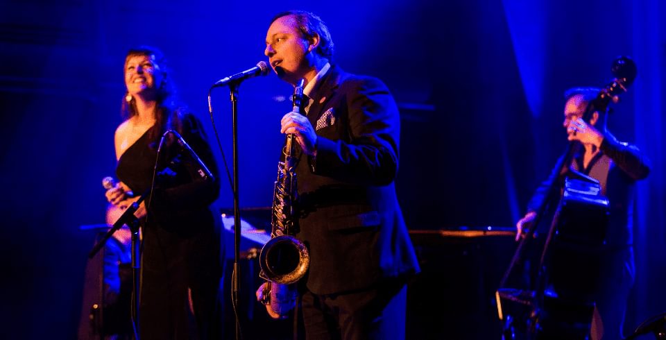 A band on stage, a singer, saxophonist and cellist in a blue light