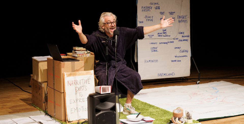 A man on stage in a grey shirt and white shorts by a stack of boxes and a green rug with a white board