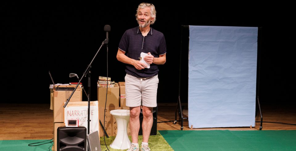 A man on stage in a grey shirt and white shorts by a stack of boxes and a green rug with a white board
