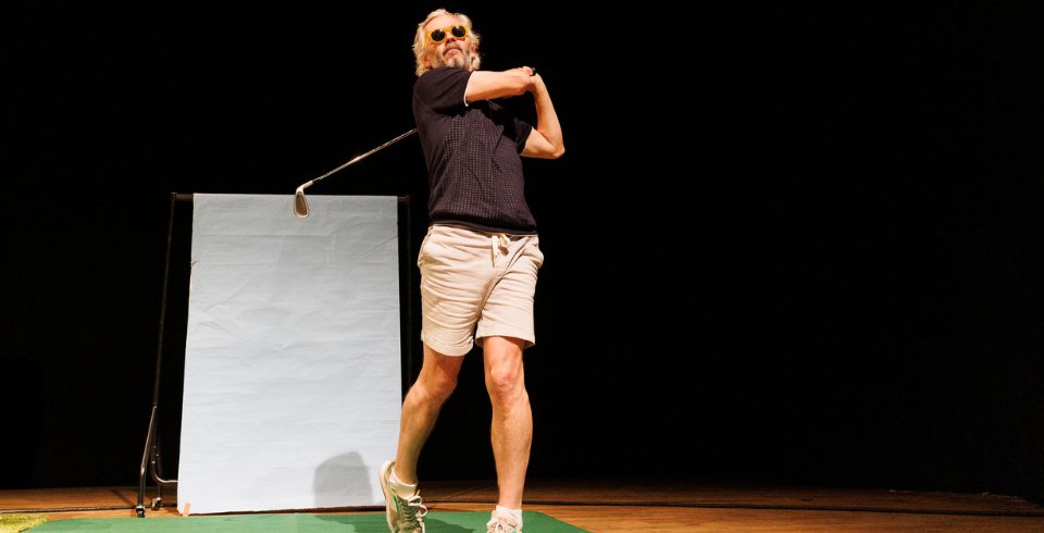 A man on stage in a grey shirt and white shorts holds a golf club with sunglasses