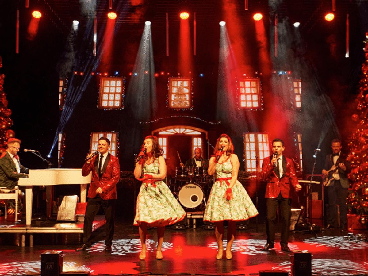 Two women with white dresses and red bows sing on the stage beside two Christmas trees and a live band
