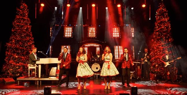 Two women with white dresses and red bows sing on the stage beside two Christmas trees and a live band