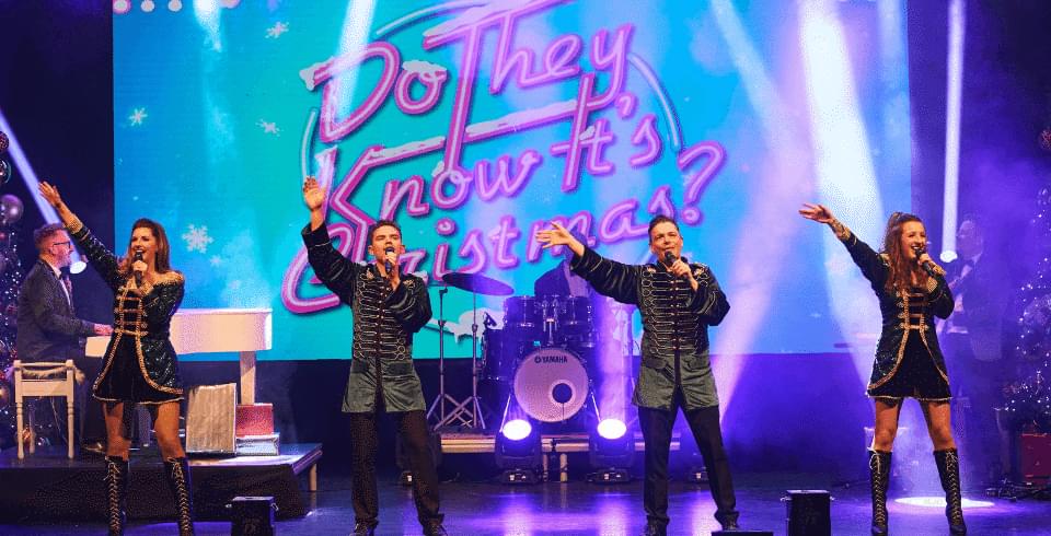 A band singing into microphones stand in glittery dark outsits in front of a blue screen that says 'Do They Know It's Christmas?'