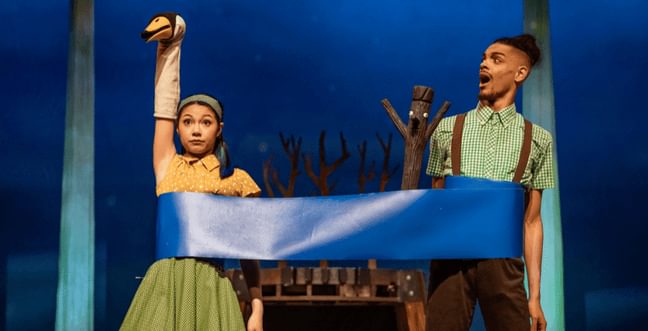 A woman with a swan puppet on her hand, stands behind a blue ribbon. This ribbon is wrapped around her and another man with a stick man puppet