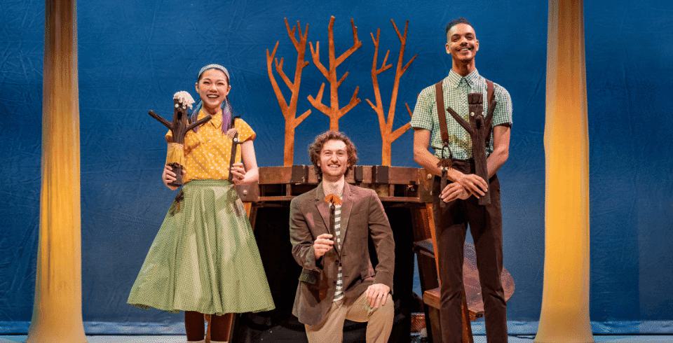 Three people on stage stand in front of a tree, all holding models of stick men