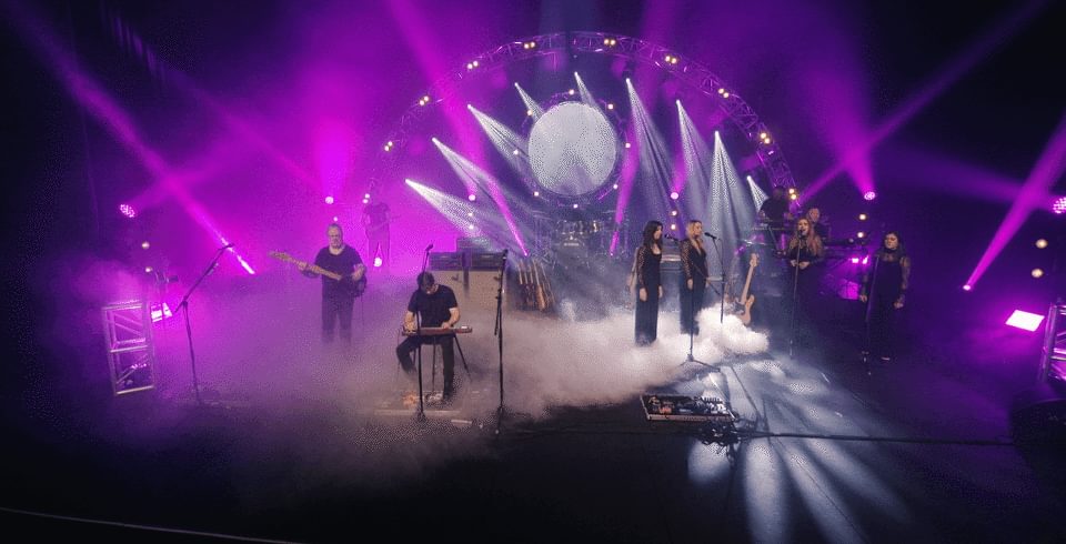 A band stand on stage with back-up singers with a large moon projection and pink beaming lights