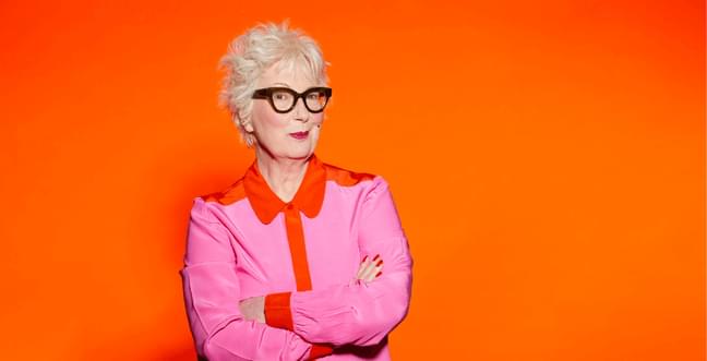 A woman with short blond hair, black framed glasses and bright red lipstick stands with crossed arms on a bright orange background.