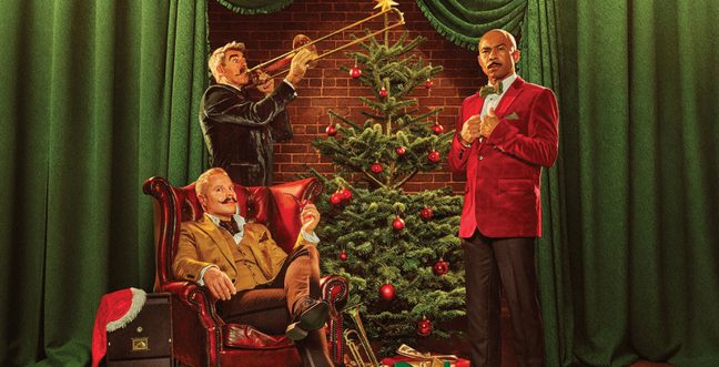 Three men in festive attire pose in front of a decorated Christmas tree with red ornaments. One man in a tan suit sits in a red armchair, another in a dark blue jacket plays a trombone, and a third in a red velvet jacket stands with his hands clasped.