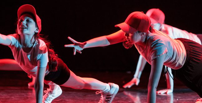 Three children on stage in red light dance with one arm outreached