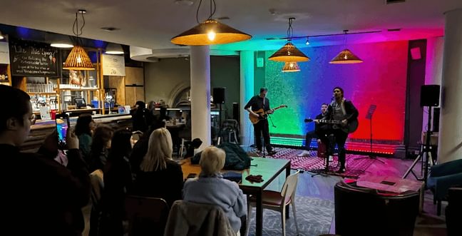 A band in front of green blue and pink gradient-lit backdrop perform to a room of people sat at wooden tables