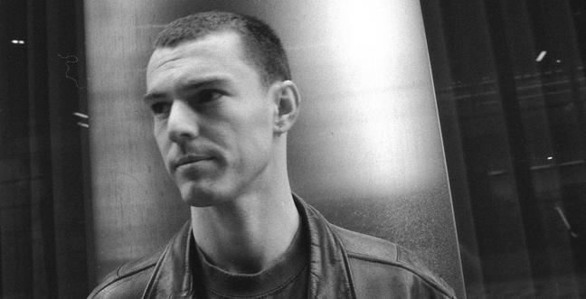A man in black and white with a shaved head stands close, looking away