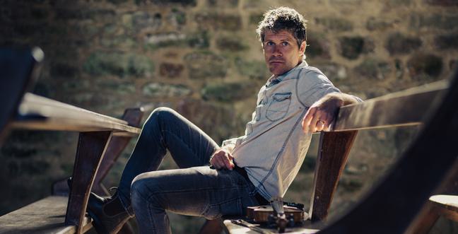 A man sits on a bench in a stoned building wearing a pale blue shirt