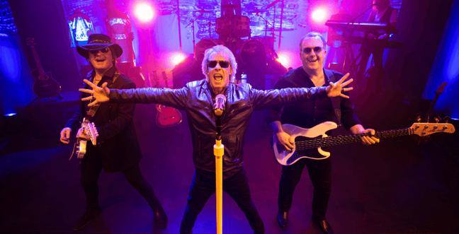 A man with blonde hair and sunglasses stands in front of two guitarists, all are dressed in black