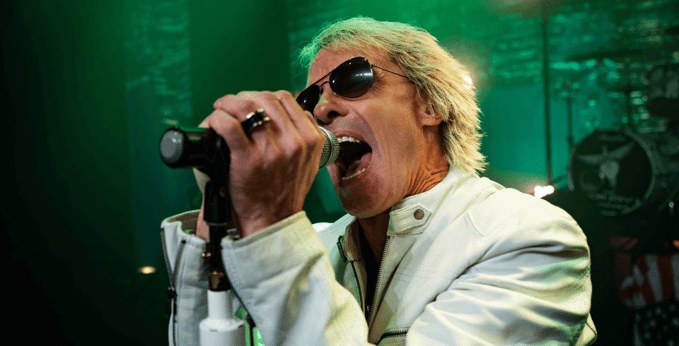 A man in a white jacket with blonde hair and sunglasses sings into a microphone