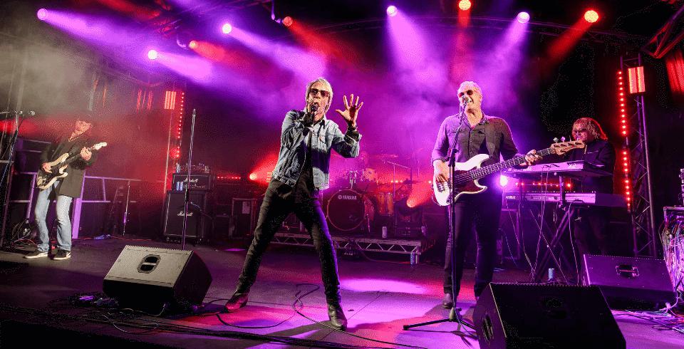 Lit by purple and red lights a man with blonde hair stands between his two guitarists as he sings