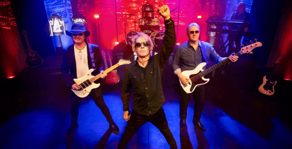 A man with blonde hair and sunglasses stands in front of two guitarists, all are dressed in black