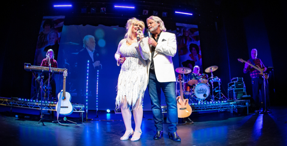 A woman with blonde hair and a white dress stands next to a man on stage singing into a microphone. He wears a white blazer and blue jeans.