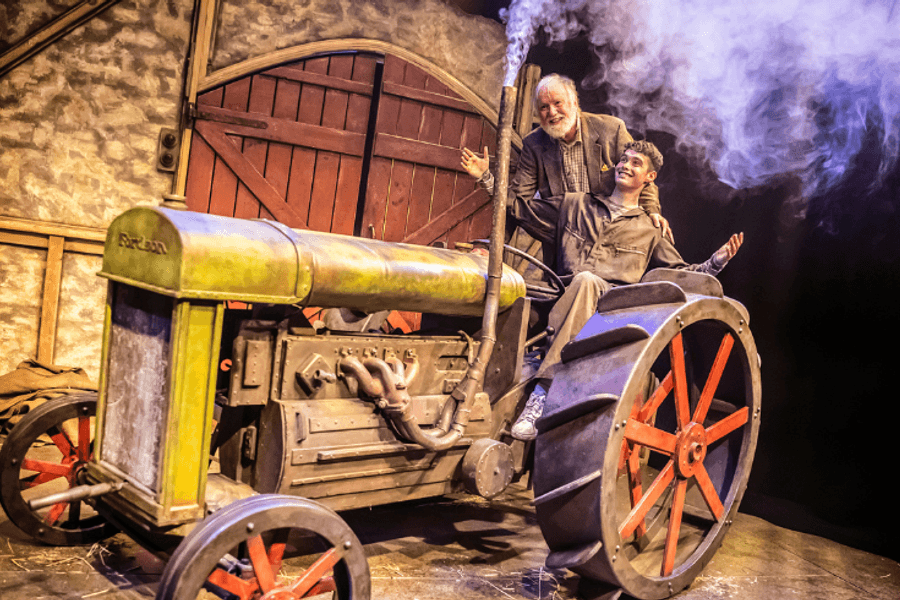 An elderly man sits on top of an old tractor which has steam coming out of its funnel