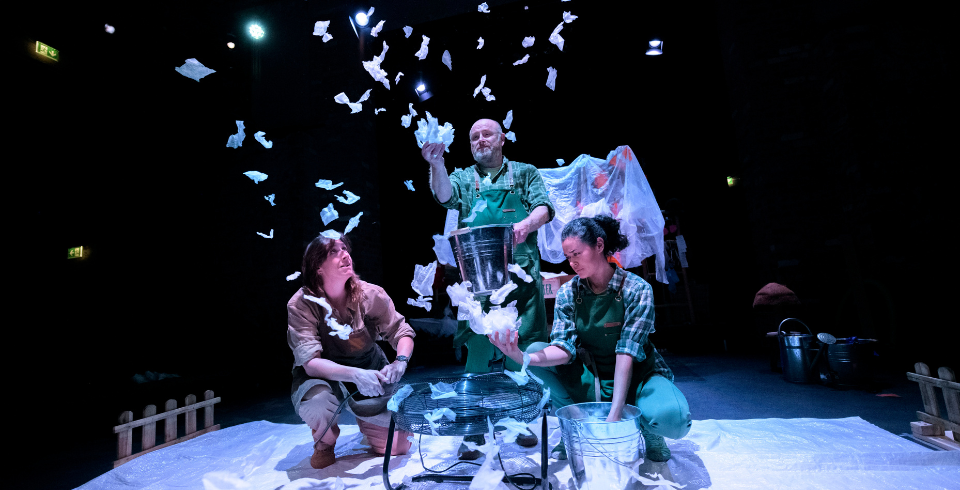 Three people on stage in green overalls stand below a bucket with white paper flying in the air