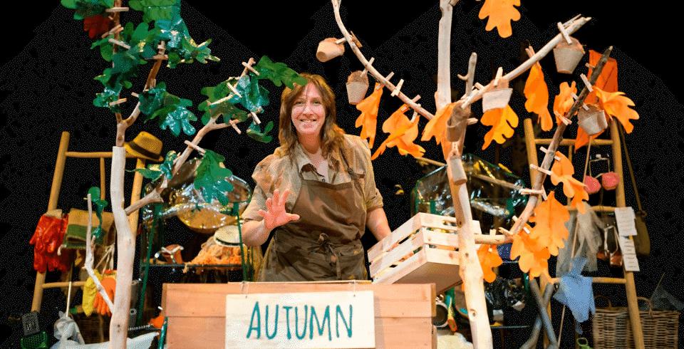 A woman stands behind two trees with green and orange leaves on stage wearing a brown shirt and apron in front of a sign that says 'Autumn'