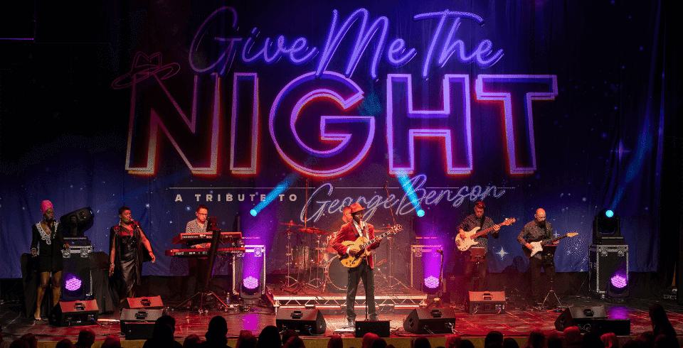A band on stage in front of the title 'George Benson Give Me The Night'
