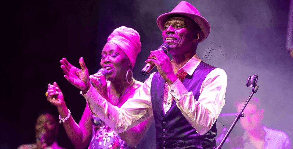 Two people on stage in pink lights sing into microphones