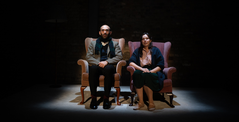 A couple sit on two armchairs in spotlights on stage, the woman in a pink shirt tucked into a long skirt with a dark cardigan and a man in a blazer, black shirt and black scarf