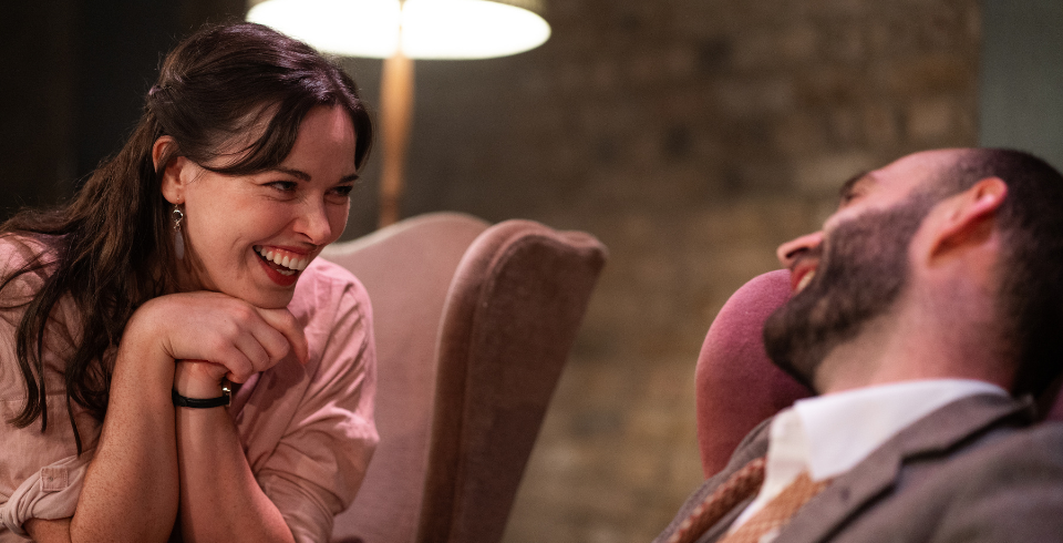A woman in a pink shirt with brown hair laughs sitting next to a man in a suit with a beard, sat on arm chairs