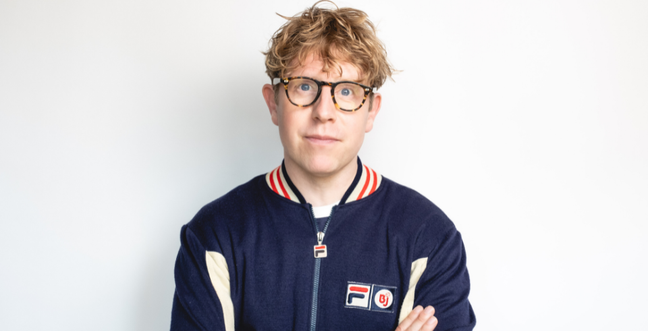 A white man with curly ginger hair stands against a white backdrop with a blue jacket, brown glasses and crossed arms