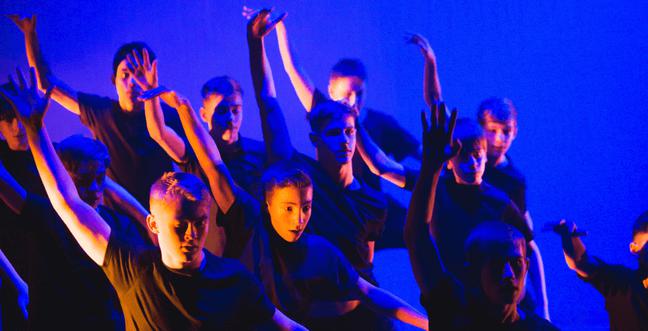 Lit by blue light a group of young male dancers jump to the right with their right arm up.