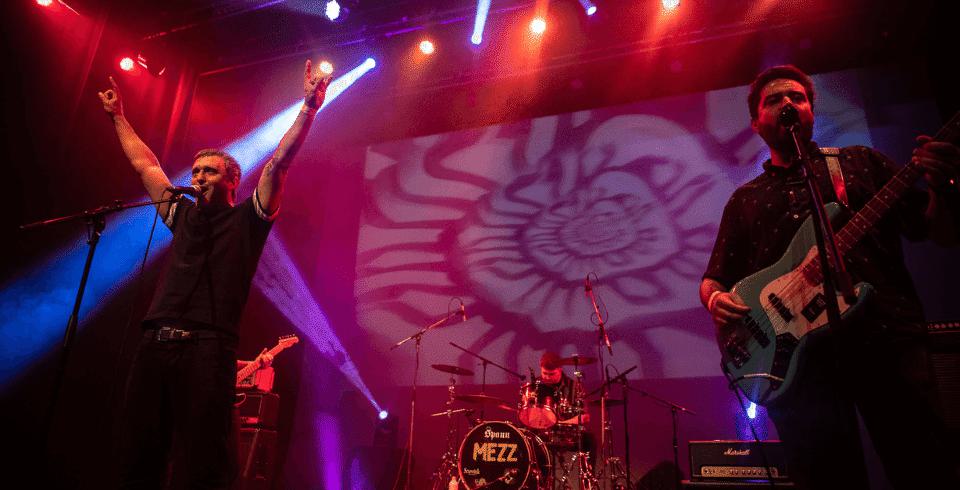 All dressed in black, the singer puts both arms up, the guitarist to his left sings while playing and between them sits the drummer at the back of the stage